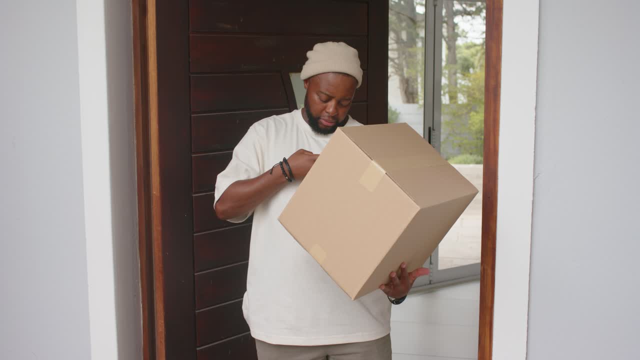 African American man pausing at front doorway holding large box, checking phone alert for delivery