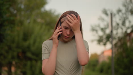 young woman appears exhausted during phone call, holding her left hand to her forehead in frustration, with a blurred green outdoor and building in background