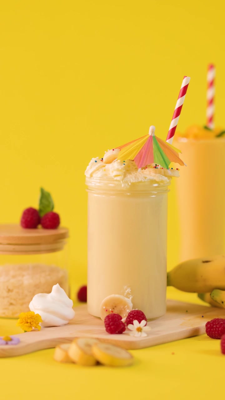 Una mano decora un cremoso smoothie alla banana con un ombrellino su sfondo giallo brillante, primo piano statico