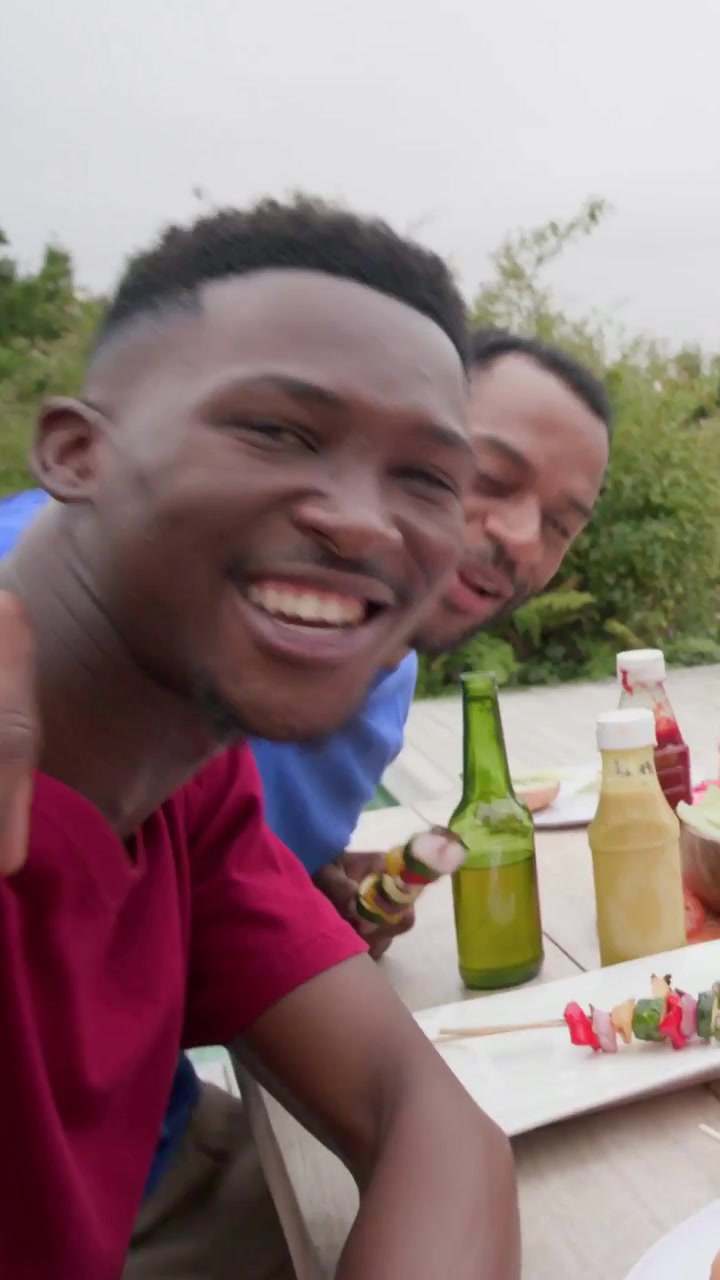 Verticale video: Drank wordt ingeschonken naar man met vlag-schildering, mannen in felgekleurde T-shirts reageren op camera op het terras.