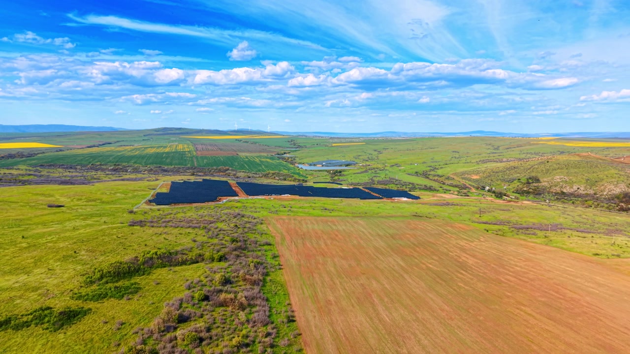 Luftfoto af et grønt landskab med en fjern solcellepark. Droneoptagelse fra høj vinkel af landbrugsmarker, et solenergianlæg til vedvarende energi, blå himmel med skyer.