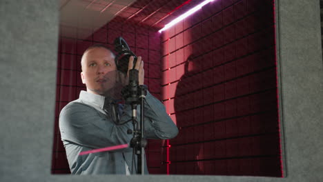 a man wearing a blue long sleeve shirt stands in front of a studio microphone, holding a headphone close to his ear in a red soundproofed room