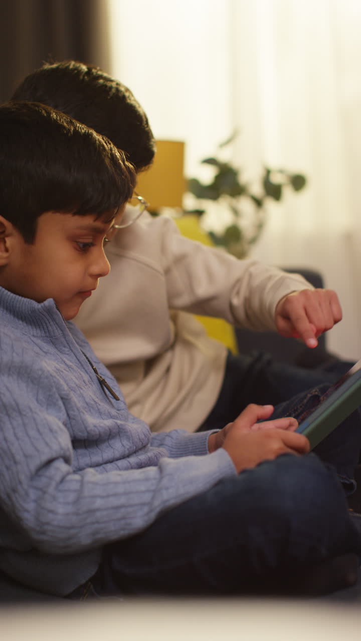 Vertical Video Of Two Young Boys Sitting On Sofa At Home Playing Games Or Streaming Onto Digital Tablet Together