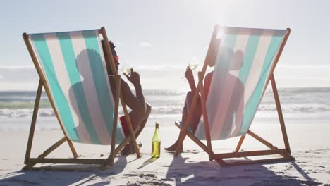 Afroamerikanisches-Paar-Trinkt-Gemeinsam-Wein-Und-Sitzt-Auf-Liegestühlen-Am-Strand