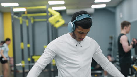 portrait view fitness enthusiast wearing over ear headphones and white zip top performing dumbbell lateral raise, focusing on form in busy gym environment with other athletes training in background