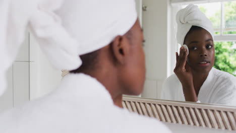 Mujer-Afroamericana-En-Bata-De-Baño-Limpiando-Su-Piel-Con-Un-Algodón-Mientras-Se-Mira-En-El-Espejo.