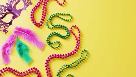 video of pink mask, mardi gras carnival beads and feathers on yellow background with copy space