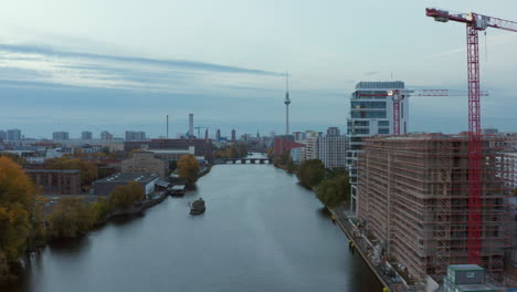 Spree-River-with-Berlin-Cityscape-and-construction-site-on-the-riverside,-Aerial-wide-angle-forward-dolly-shot