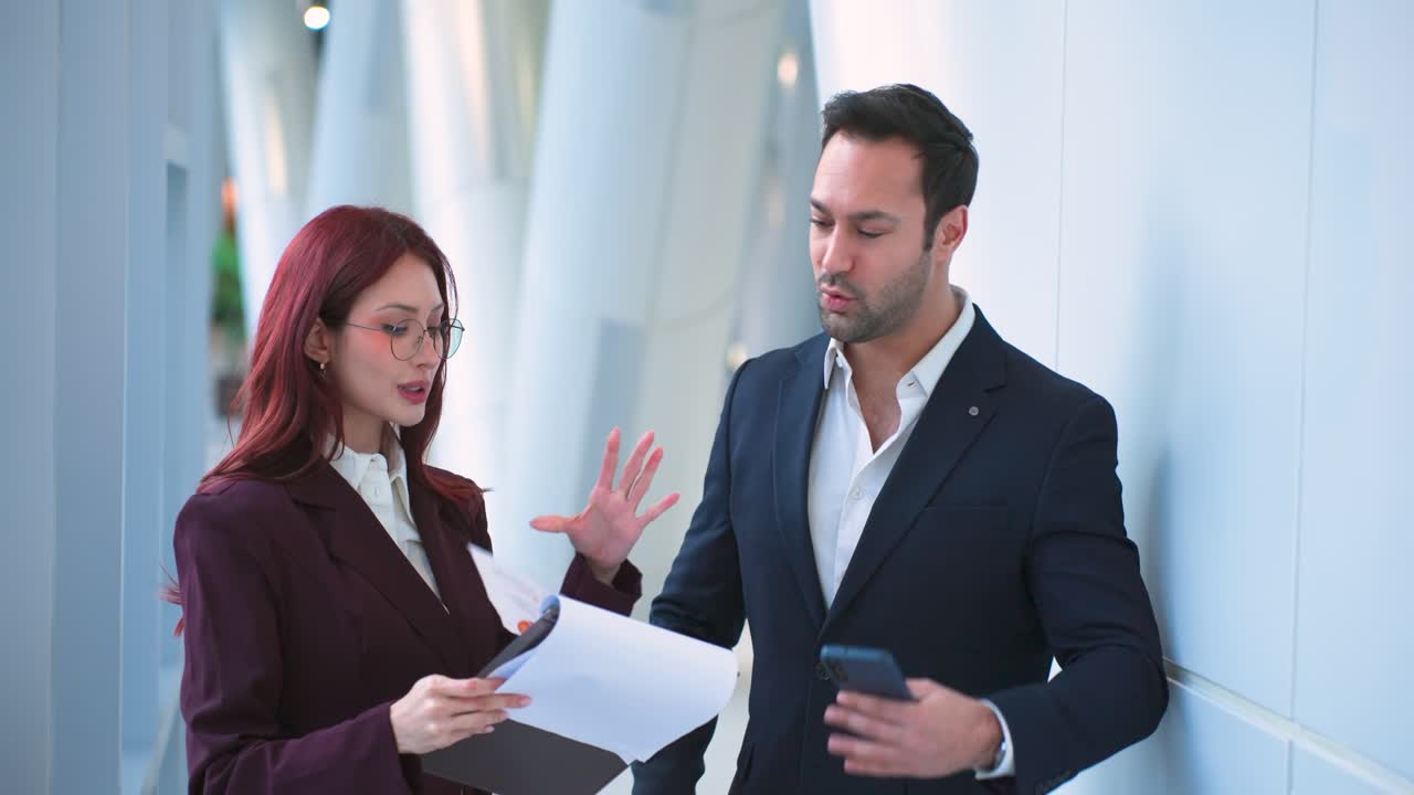 Mulher latina profissional de terno cor vinho e homem europeu de blazer azul-marinho celebrando o sucesso com um high-five enquanto ele fala ao telefone segurando documentos em um corredor de escritório moderno.