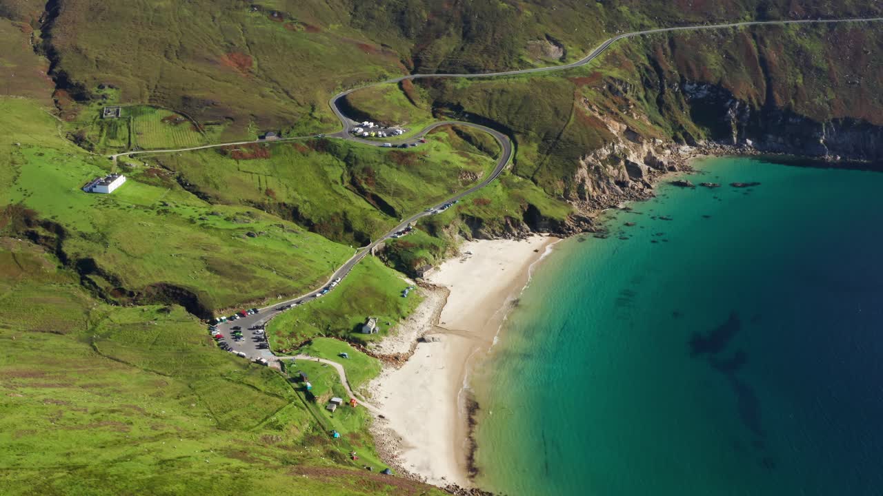 Aerial orbit of Keem Beach with calm blue Atlantic waters, cars parked by the coastline, and a winding mountain road in County Mayo