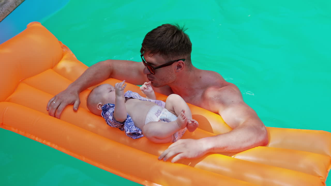 Caucasian man standing in the swimming pool. Tiny infant boy lies on the inflatable mattress crying. Top view.