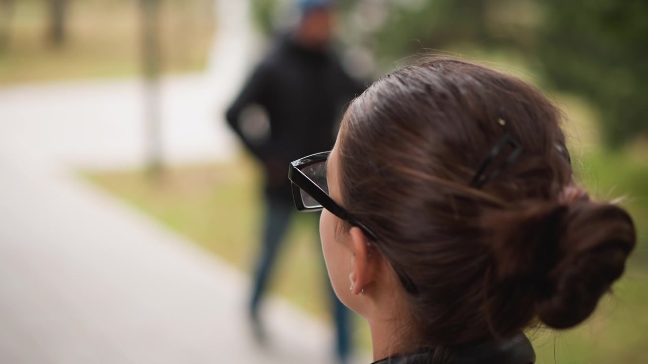 Close up of woman with glasses observing another woman walking away on path in outdoor park setting. scene features soft natural light and shallow depth of field emphasizing back of her head