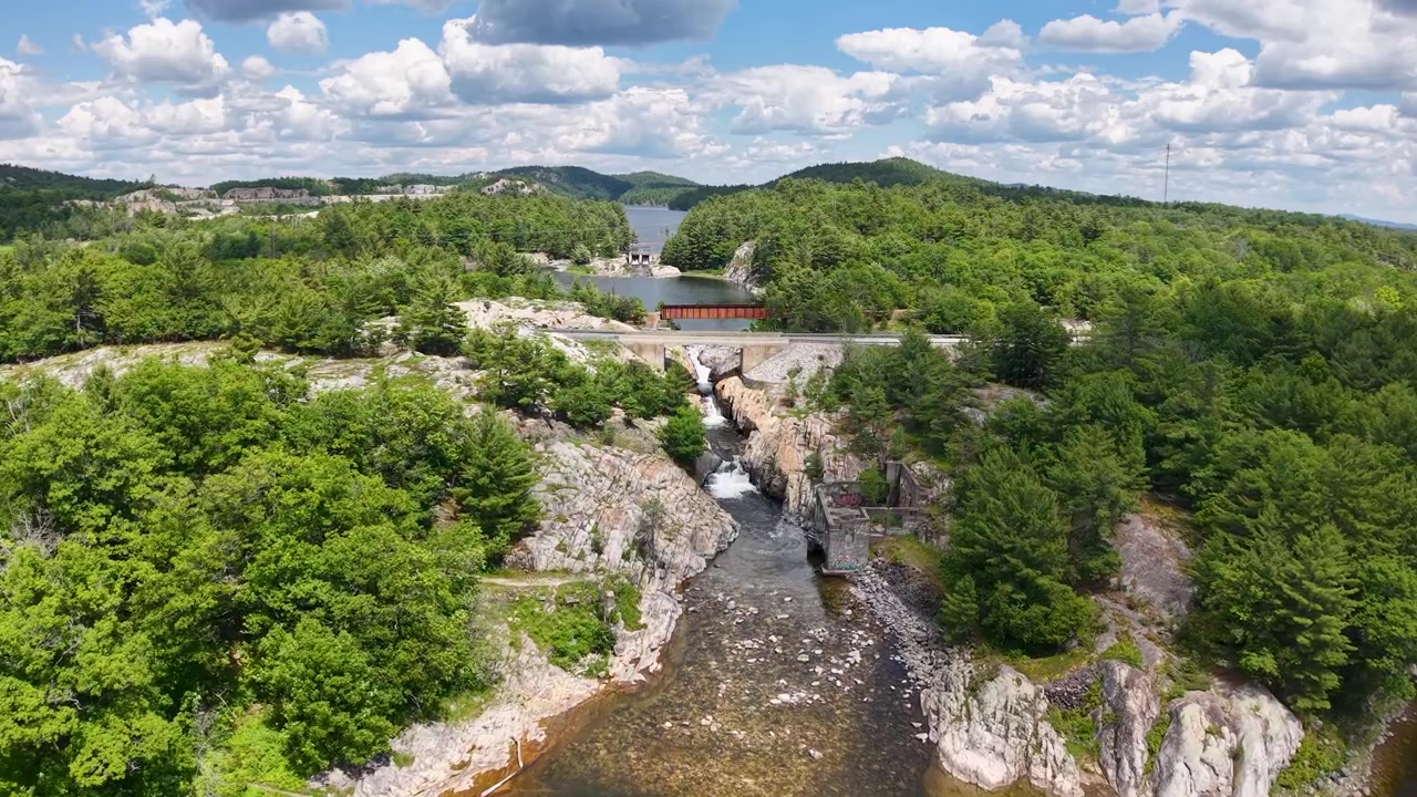 High drone view looks upstream through a rocky forest gorge toward a road bridge crossing above the Whitefish Falls river