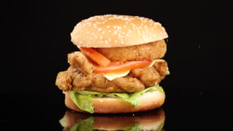 golden french fries fall beside a stacked fried chicken burger with vegetables and onion ring, shot in slow motion against a black background with studio lighting