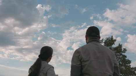 rear view of woman pointing at distant hot air balloon drifting across cloud-filled sky while man watches beside her, surrounded by trees and soft morning light