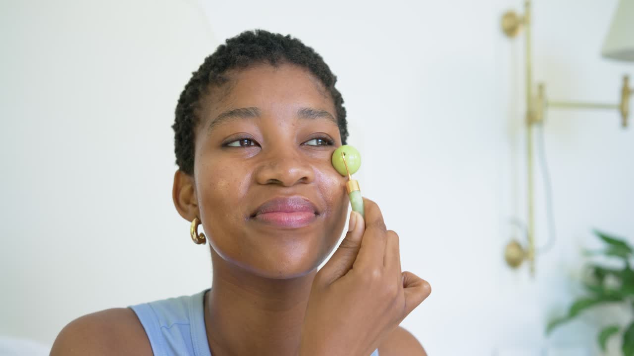 Mulher adulta africana segurando um rolo de jade com pele luminosa no quarto, massageando para reduzir o inchaço.