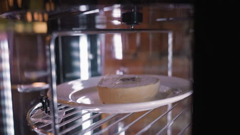 Creamy dessert cake on white plate rotating inside display fridge, captured in soft lighting with reflections on glass and metallic surfaces enhancing texture and presentation of chilled treat