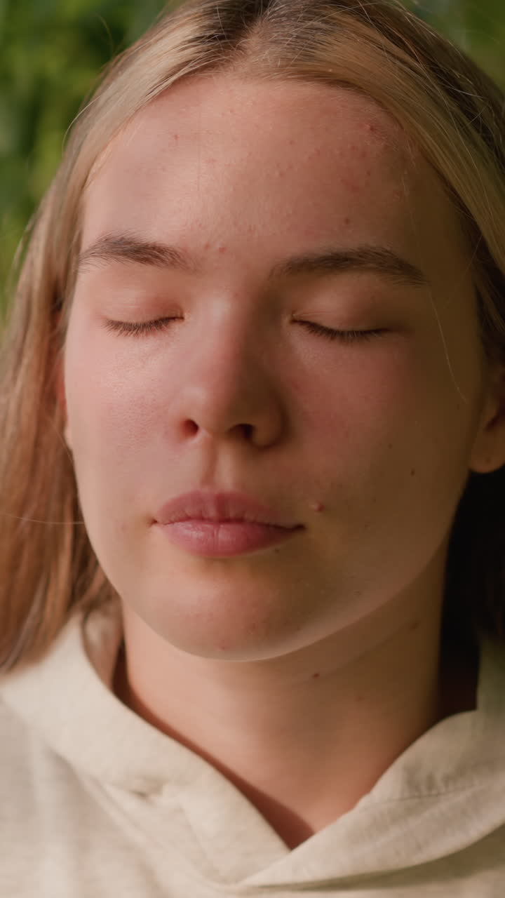 Close-up of young lady closing and opening her eyes, basking in gentle sunlight with serene expression, enjoying soft breeze in natural setting, blurred green background with swaying leaves
