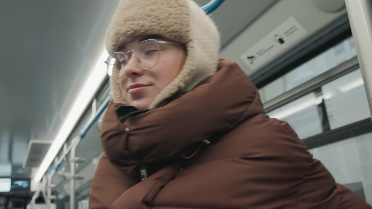Passenger bundled in brown puffer coat and fuzzy earflap hat moves forward down public transit aisle while holding overhead handrail, showing steady stride, slight lean, and routine winter commute