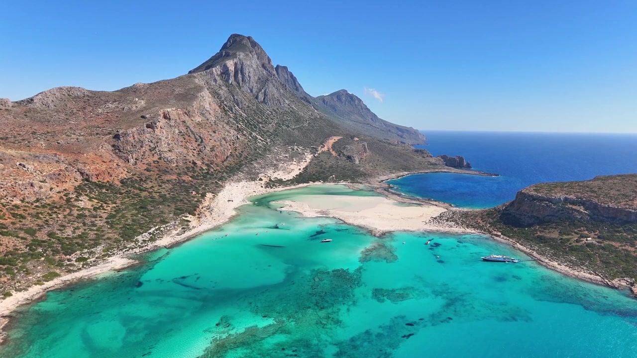 Balos-lagunen udvider sig under luftkameraet, indrammet af levende turkise lavvandede områder, blegt sand, et barskt bjerg og dybblåt vand langs den kretensiske kyst