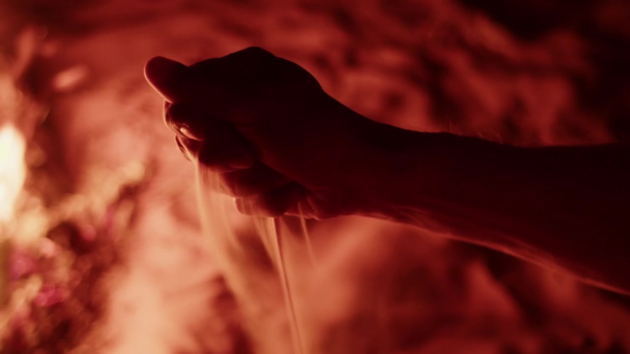 Close-up de uma mão derramando areia fina do Saara à noite na Tunísia. Cena desértica atmosférica com iluminação dramática, focando na textura dos grãos de areia e no conceito de tempo ou viagem