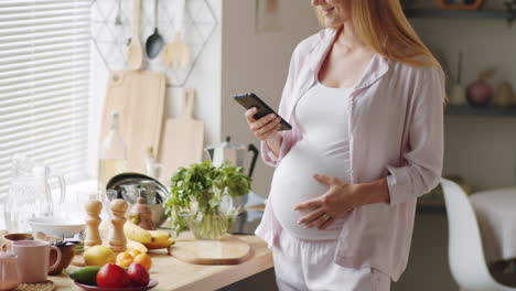 pregnant woman messaging on smartphone in kitchen