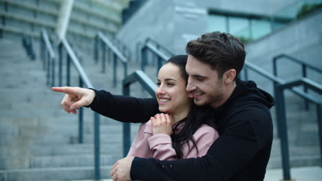 Portrait-of-smiling-couple-hugging-outdoor.-Cheerful-man-pointing-finger-to-side