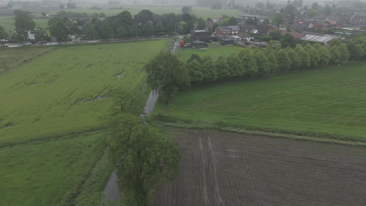 Drone shot in the rain in Belgium of a group of cyclist biking in a swarm during a race in between the trees on a grey and wet day LOG