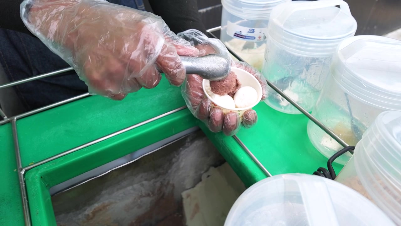 Close-up of a vendor in plastic gloves serving ice cream scoops into a cup.
