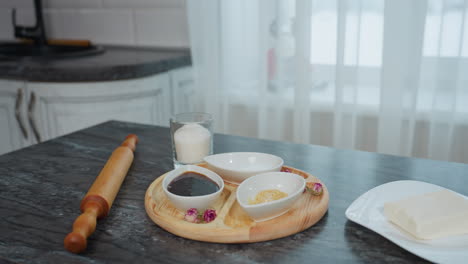 honey in plates on wooden tray with kitchen essentials, sugar, butter, and rolling pin on sleek black table, white curtain allows soft natural light, revealing snowy building through window