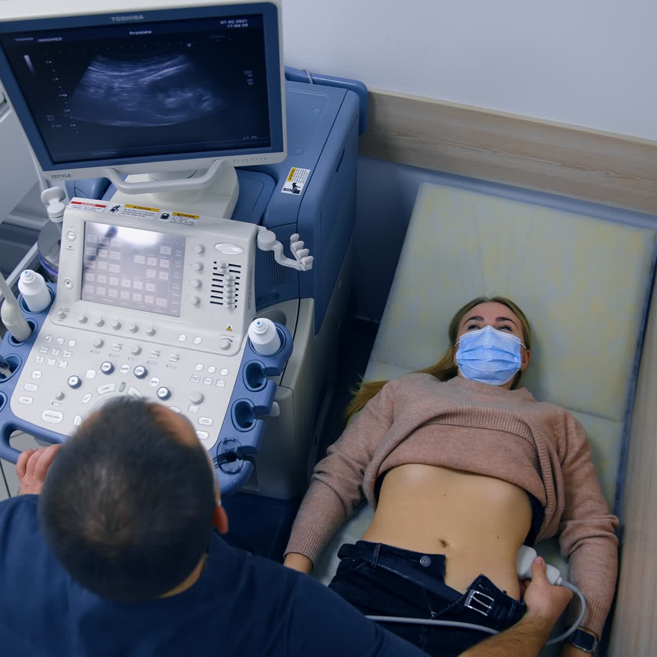 Blonde woman wearing mask lies on the doctor's couch. Male obstetrician uses ultrasonic device for patient's health examination. Top view