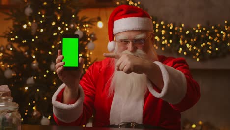 santa points his finger at the green screen mobile phone screen. christmas sale. elderly santa claus shows a screen with a chromakey