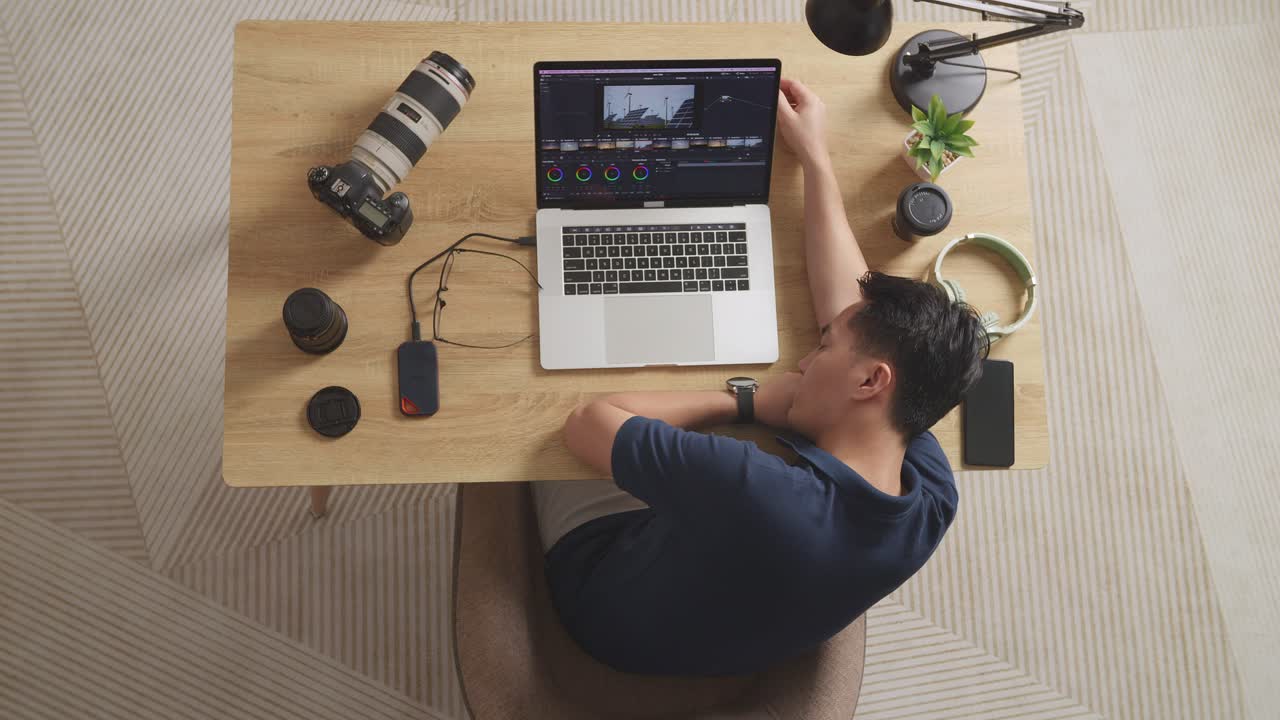 Top View Of Asian Male Color Grading Sleeping While Sitting In The Workspace Using A Laptop Next To The Camera Editing The Video At Home