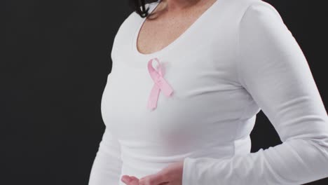 Mid-section-of-a-woman-showing-the-pink-ribbon-on-her-chest-against-black-background