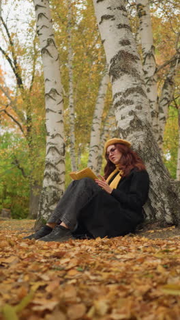 tourist enjoying book in solitude wears yellow beret scarf around neck, deeply focused on book in beautiful autumn park, surrounded by golden leaves and birch trees