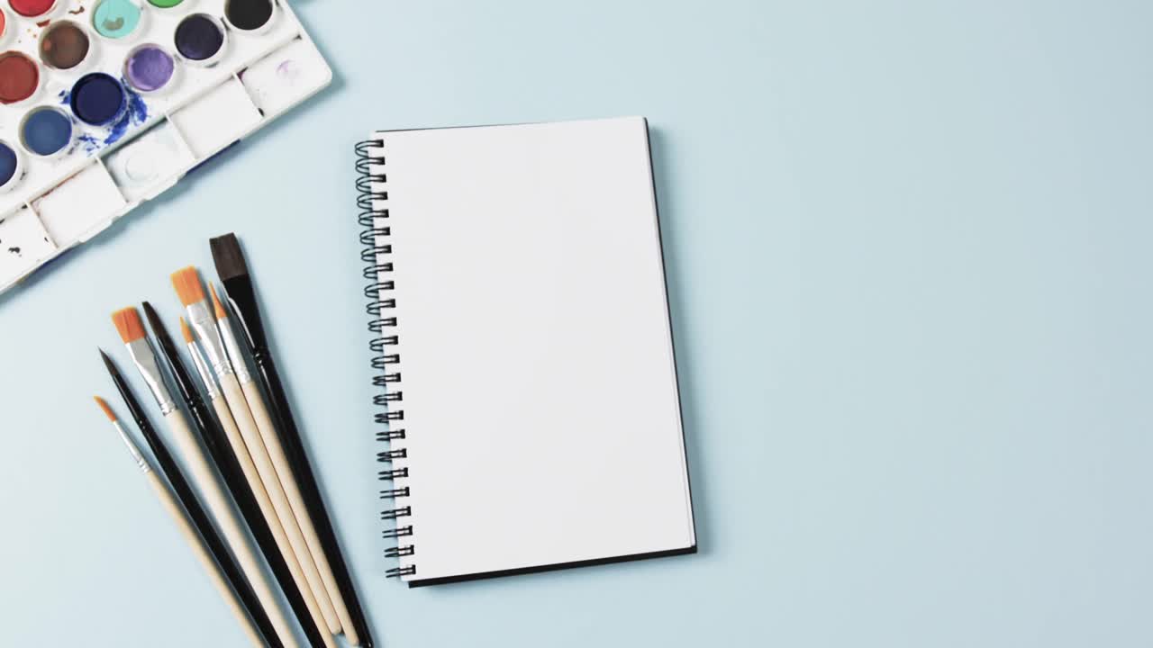 vista aérea del cuaderno, los pinceles y las pinturas sobre fondo azul con espacio de copia, en cámara lenta