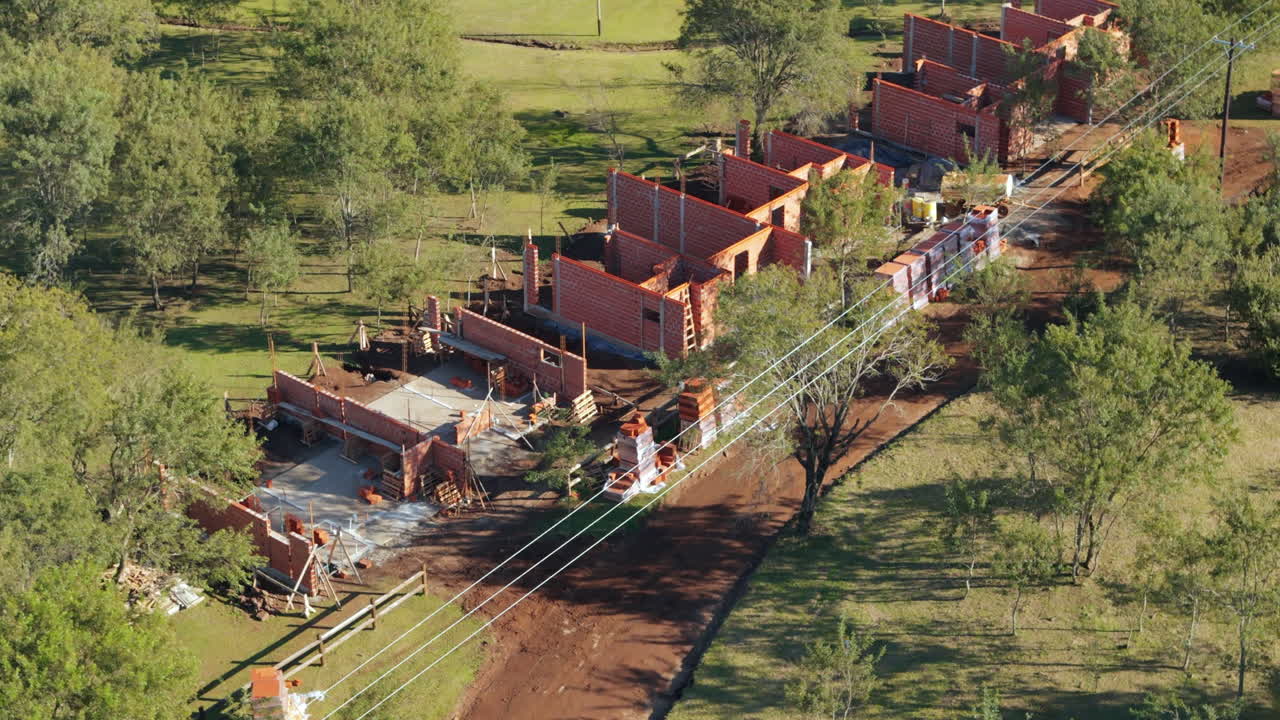 Drone reveals a construction site with several red brick buildings in early stages of development surrounded by greenery and rural terrain, scaffolding and equipment visible, real time, reveal shot