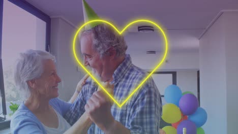 Gelbe-Neon-Herz-Ikone-Vor-Kaukasischem-Seniorenpaar,-Das-Während-Der-Geburtstagsfeier-Zusammen-Tanzt