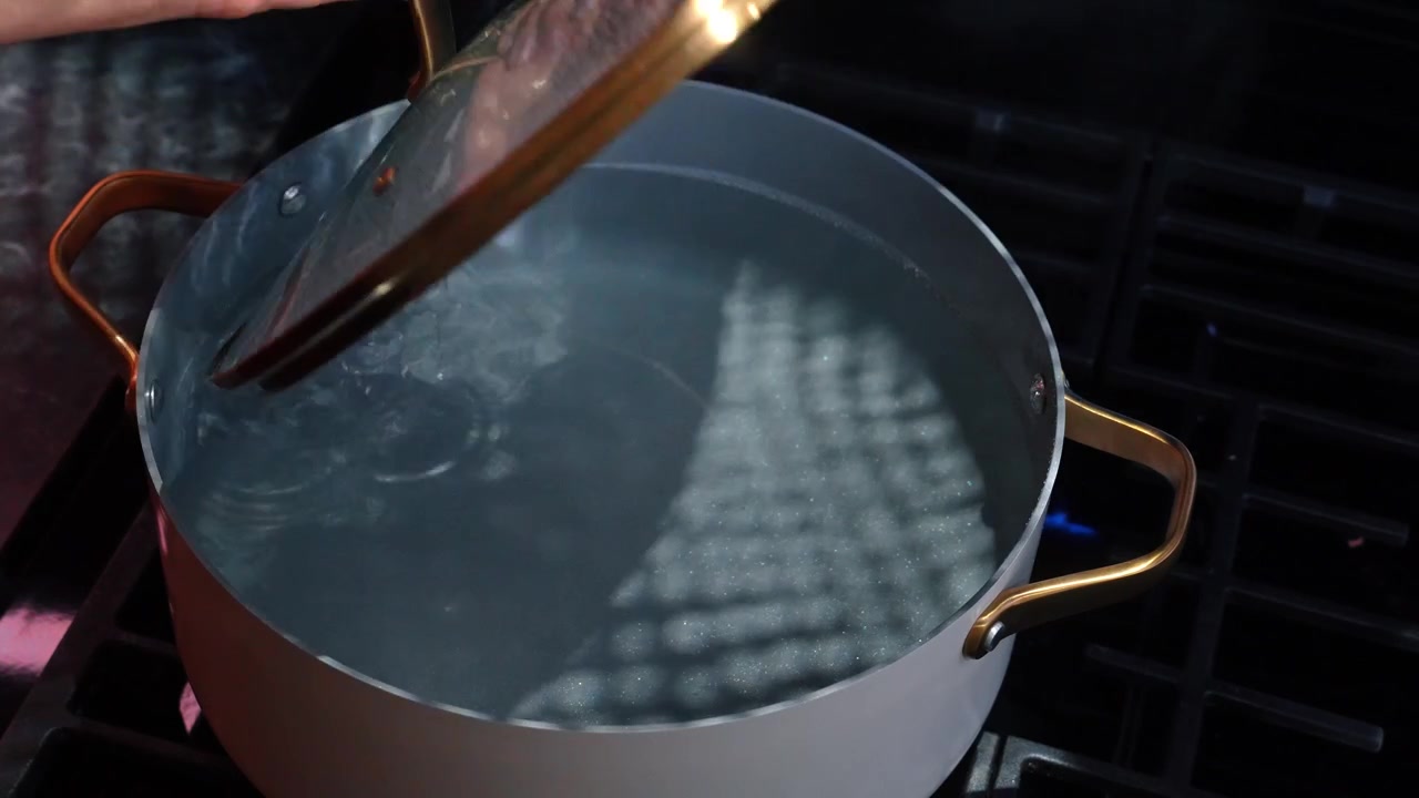 Cooking water heats in a pot on a stove with a lid being lifted. A pot sits on a stove with water inside.