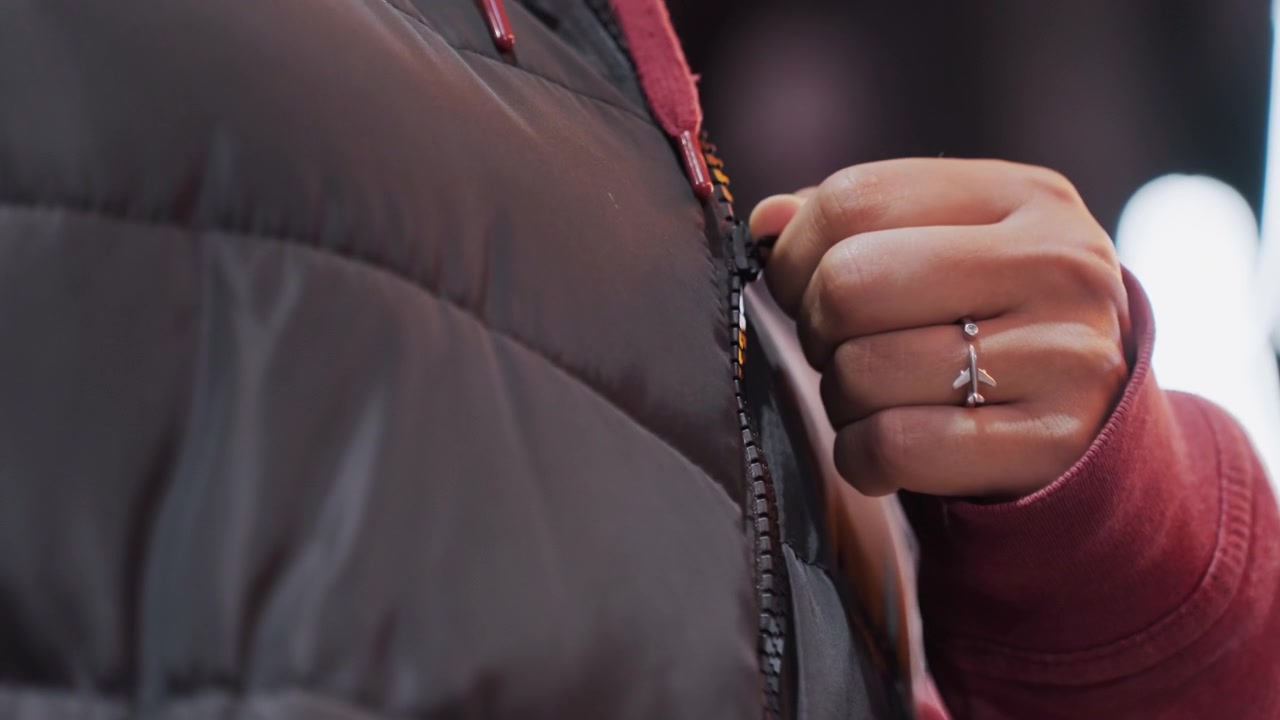 Night street fingertip securing puffer, tight framing on zipper and ring with blurred city lights, small gesture during evening walk conveying intimate urban texture and steady nocturnal pace