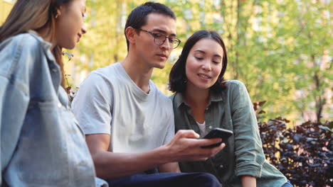 Joven-Japonés-Sosteniendo-Un-Teléfono-Móvil-Y-Tomando-Una-Divertida-Foto-De-Selfie-Con-Sus-Dos-Amigas-Mientras-Se-Sienta-Al-Aire-Libre-En-El-Parque