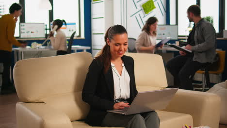 Happy-hispanic-lady-sitting-on-couch-in-start-up-business-office-typing-on-laptop