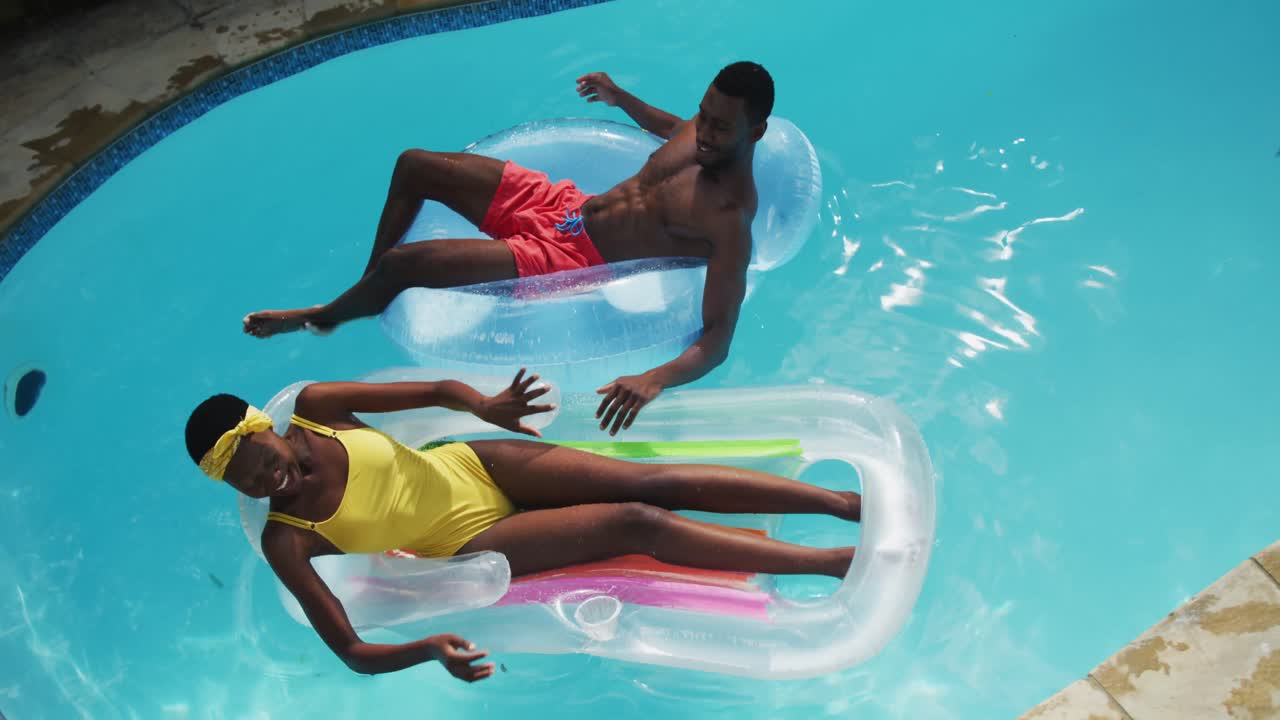 Happy african american couple lying on inflatables in swimming pool splashing each other and smiling