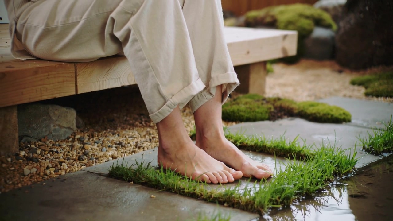 Resting on bench, adult's lower legs in trousers flexing bare feet on grass-topped stone by pond