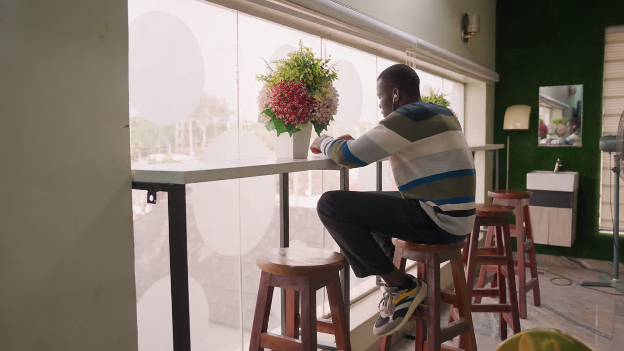 Uomo che controlla il telefono al bancone della finestra. Uomo seduto con maglione a righe che tocca il telefono, bouquet nelle vicinanze, retroilluminazione soffusa della tenda a fantasia, sorriso sottile dopo un messaggio in arrivo, interni moderni con parete verde, atmosfera calda.