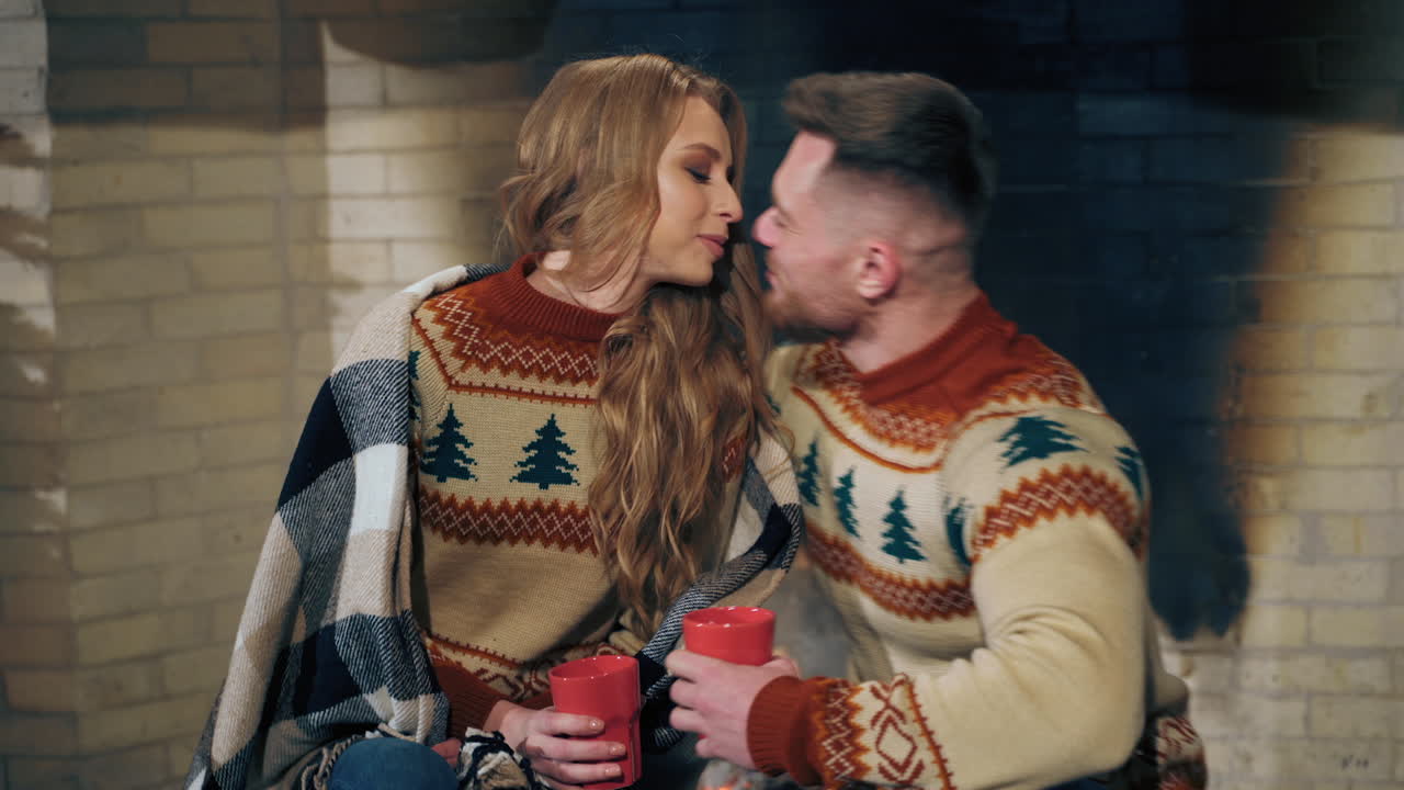Happy young man and woman kiss near the fireplace. Romantic couple in Christmas sweaters enjoying evening together in cozy home place.