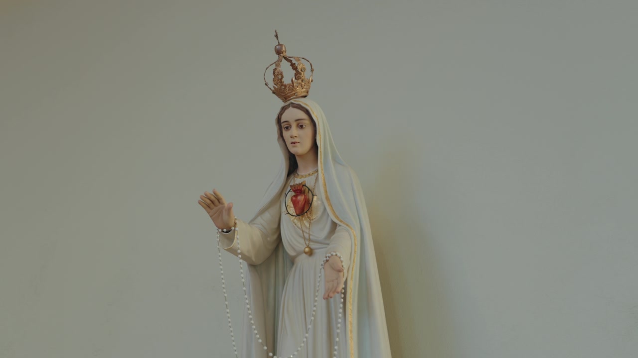 Statua della Madonna di Fatima con corona dorata, cuore rosso e rosario bianco su sfondo neutro