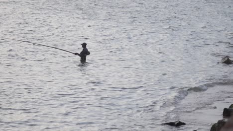 Fisherman-standing-hip-high-deep-in-the-sea-holding-fishing-rod-action-silhouette-scenic-evening-time-slow-motion-b-roll-clip,-Galle-Dutch-Fort,-Sri-Lanka