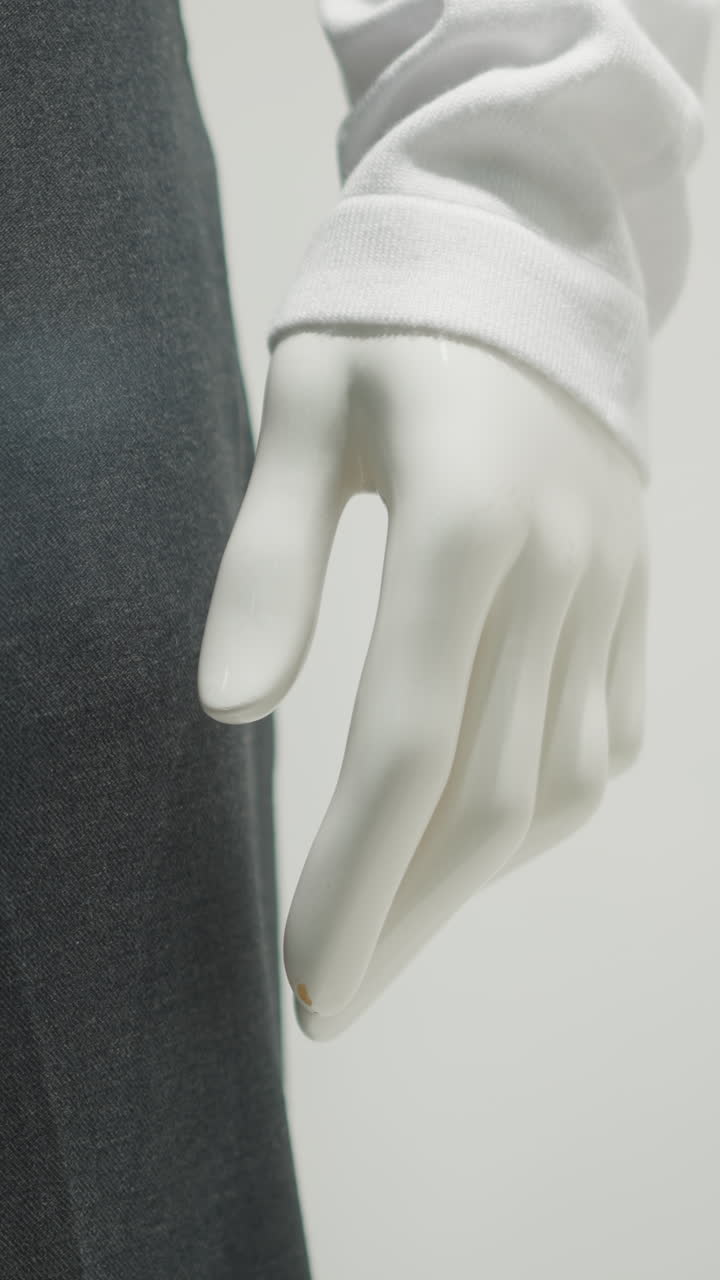 Partial view of mannequin hand wearing long white sleeve and black trousers, showcasing elegant clothing detail with soft lighting, minimalist composition, and clean background