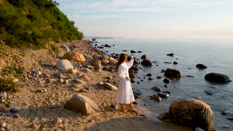 aerial approaching to pretty lady in white sundress observing colorful sunrise over baltic sea standing alone on rocky beach seaside